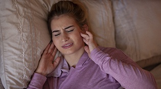 A young female lying in bed and holding both sides of her jaw in pain because of bruxism